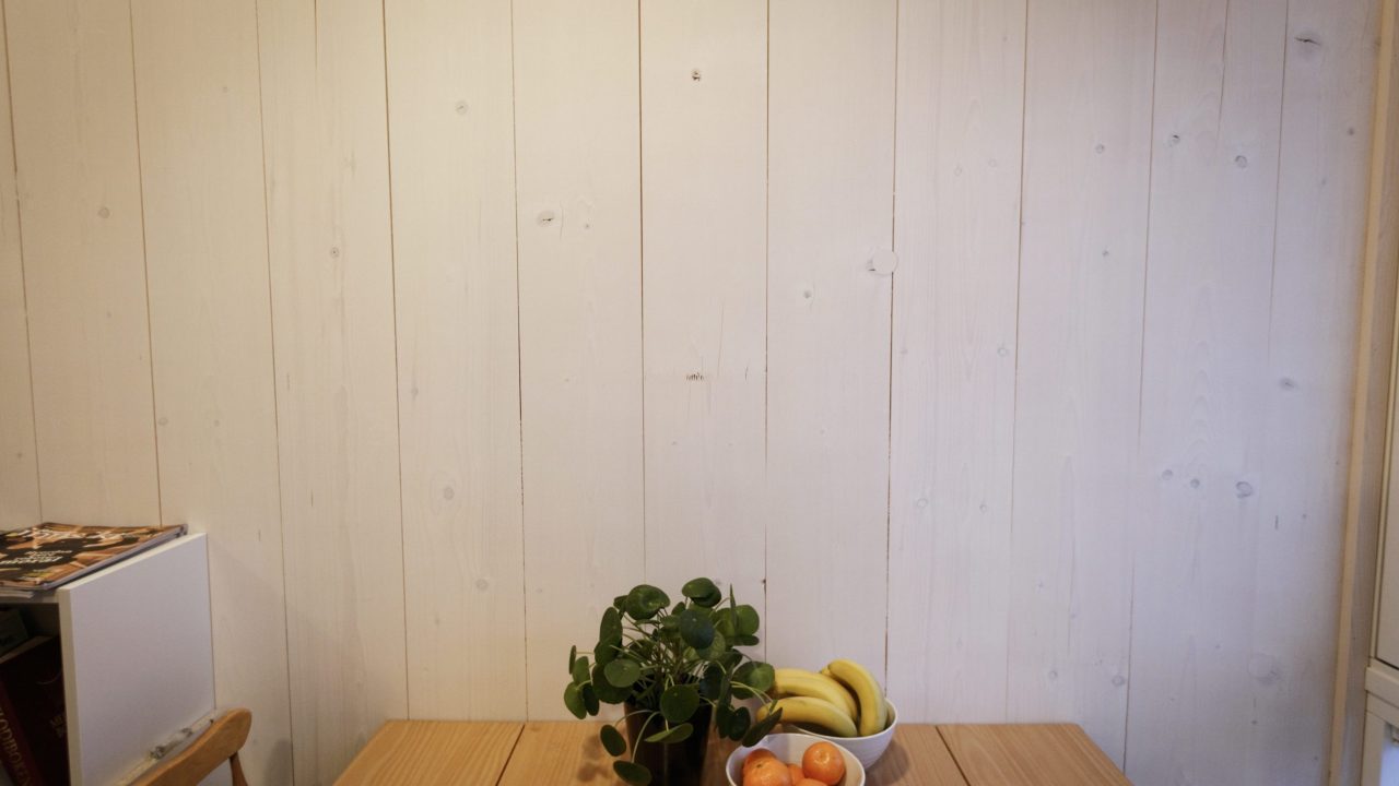 Vegg i lyst tre. Foran står et eikebord med en grønn plante og skåler med bananer og klementiner