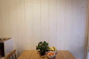 Vegg i lyst tre. Foran står et eikebord med en grønn plante og skåler med bananer og klementiner