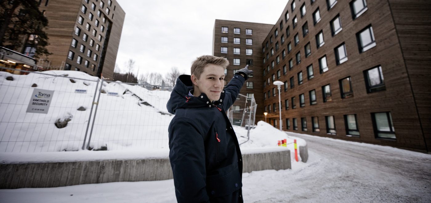 Jon Martin Høie (20) utenfor studenthuset på Kringsjå. Bygget er brunt og i massivtre. Han peker opp på leiligheten sin.
