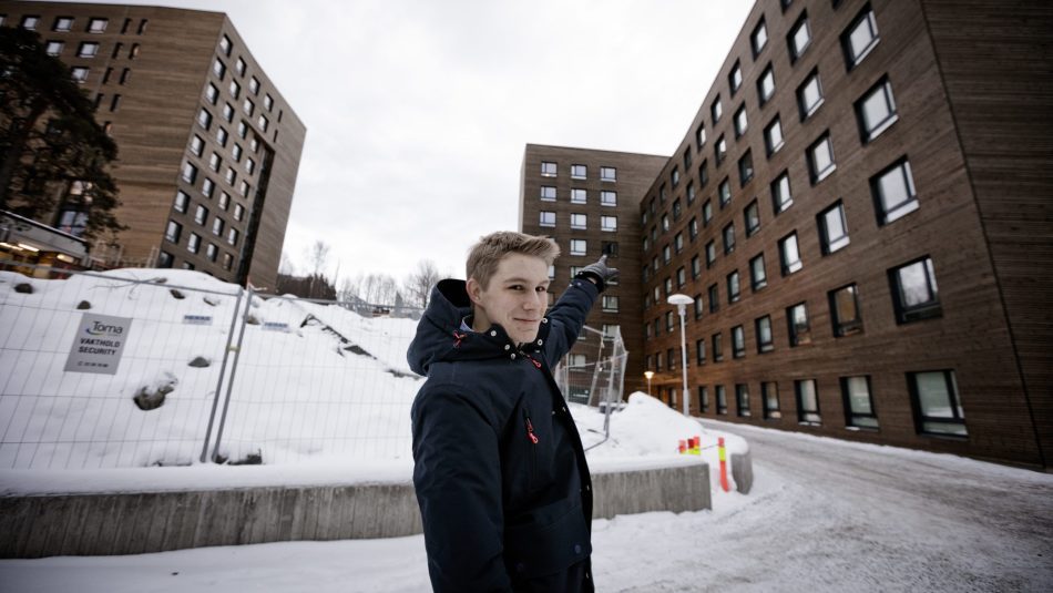 Jon Martin Høie (20) utenfor studenthuset på Kringsjå. Bygget er brunt og i massivtre. Han peker opp på leiligheten sin.