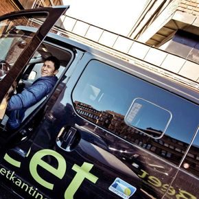 Fornøyd sjåfør sitter bak rattet på en sort elvarebil. Har åpen dør og smiler til kamera.