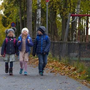 Tre barn på vei til skolen på høsten. Asfalt og gulgrønne trær rundt.
