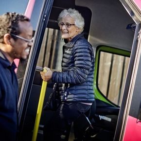 Eldre dame på vei inn i rosa buss, bussjåfør i forkant