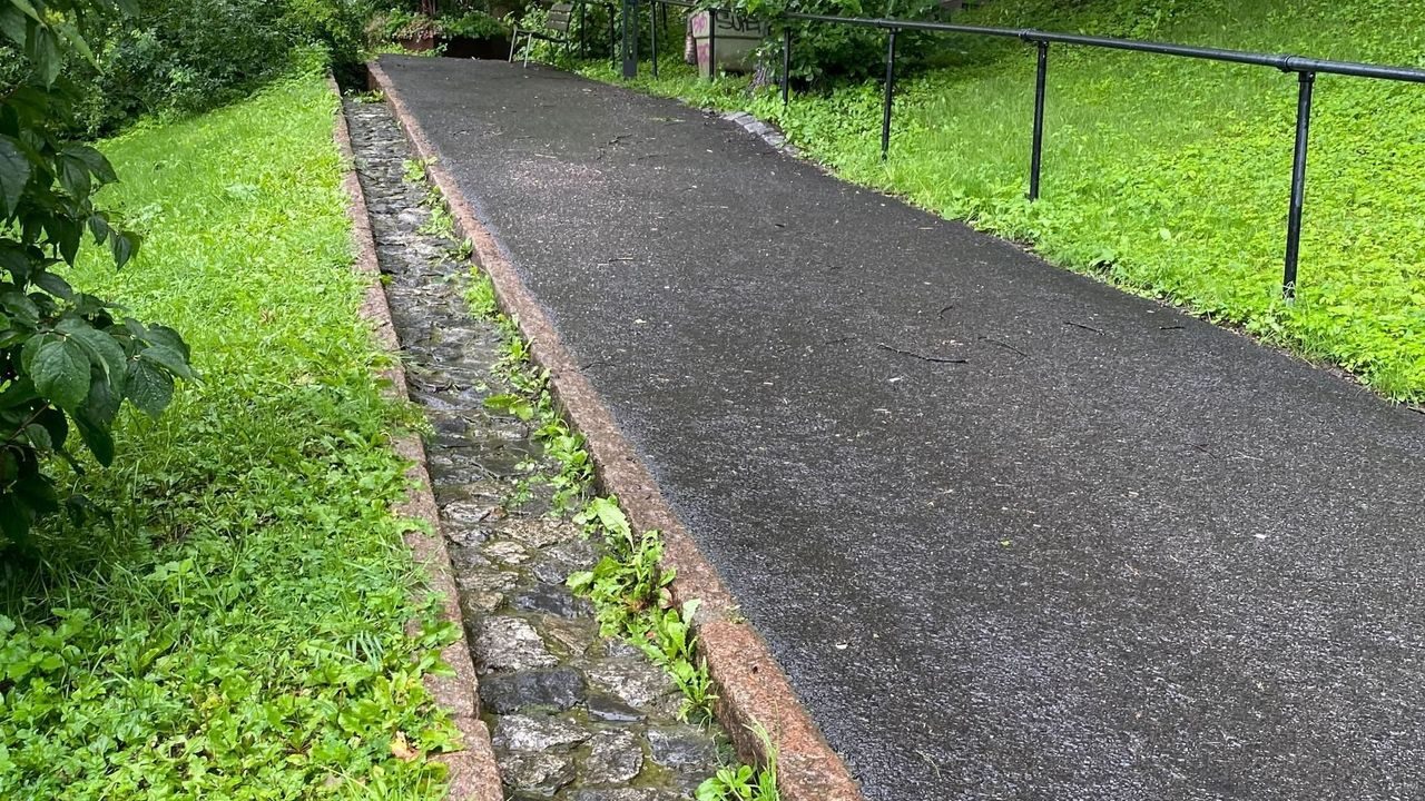 Et vanlig asfaltert gangvei i en nedoverbakke, ved siden av ser man en liten renne av stein som skal lede vannet ned i Iladalen park.
